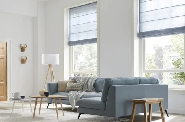 Licht en ruim ingerichte woonkamer met grote ramen voorzien van elegante, blauwe vouwgordijnen die zorgen voor een zachte filtering van het daglicht en een warme sfeer creëren.