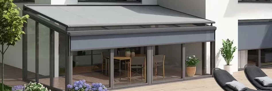 Moderne serre met zonwering en glazen schuifdeuren, omgeven door een groene tuin met bloemen en ligstoelen op het terras