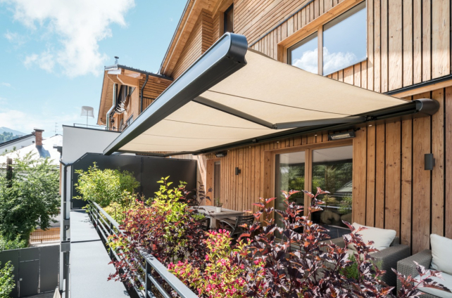 Uitschuifbaar knikarmscherm op een houten gevel, schaduw biedend aan een modern balkon met planten.