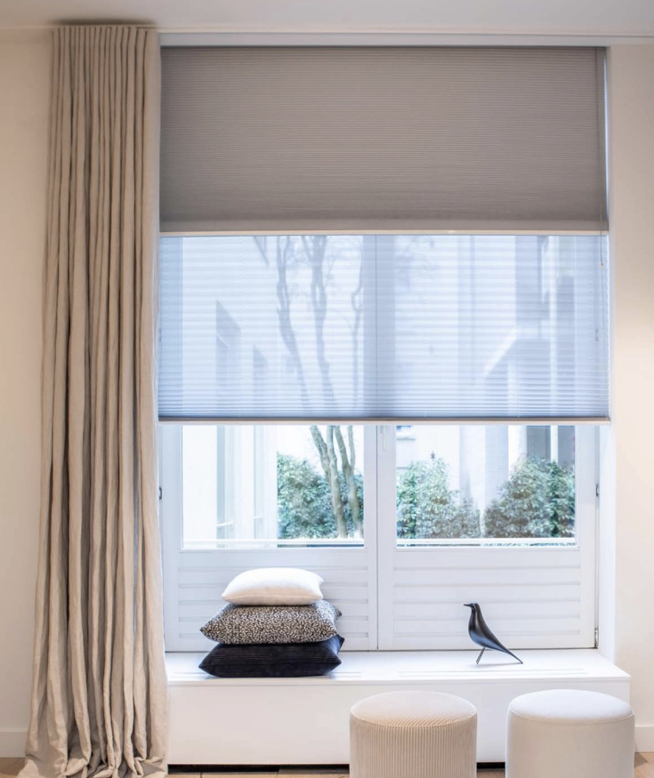 Gezellige woonkamer met een groot raam voorzien van een lichtgrijs Kvadrat rolgordijn en transparant geplisseerd gordijn, omringd door een lange beige gordijnstof en een knusse zithoek met kussens en poefs.