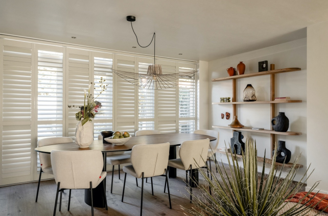 Eetkamer met witte shutters, een grote houten eettafel, beige stoelen en een minimalistisch wandrek met decoratieve objecten.