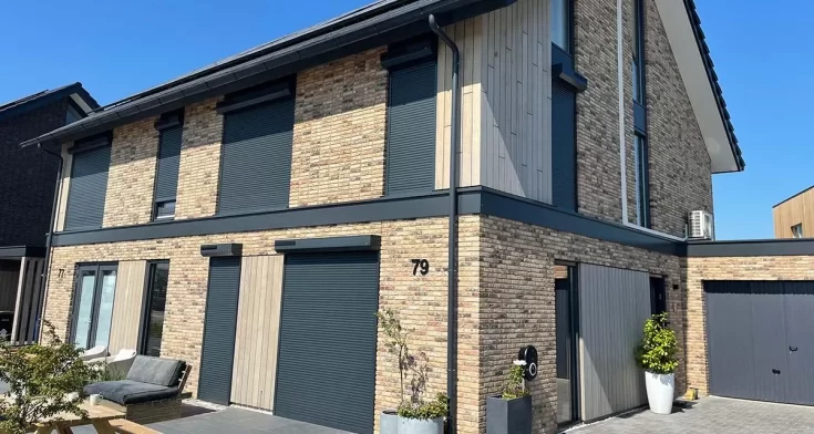 Moderne twee-onder-een-kapwoning met houten en bakstenen gevel, uitgerust met gesloten rolluiken en een strak aangelegd terras met tuinmeubelen