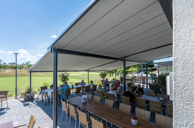 Ruime pergola met zonwering biedt schaduw aan een terras vol zitplaatsen bij een golfbaan