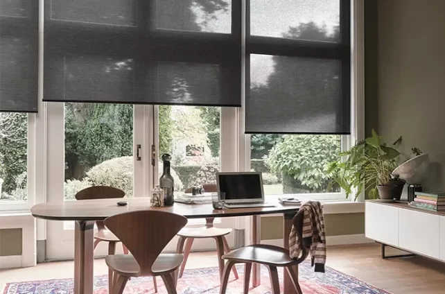 Binnen interieur met een eettafel en stoelen op een roodblauw kleed en donkergrijze binnenzonwering voor de ramen