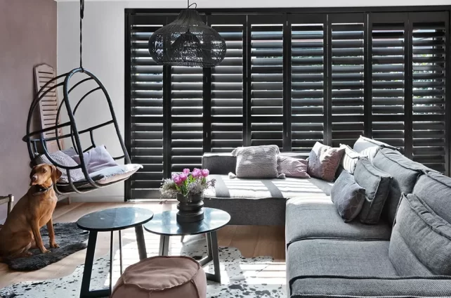 Woonruimte met een krijze l-vormige bank, een hangstoelm ronder bijzettafels en donkerbruine shutters voor de ramen