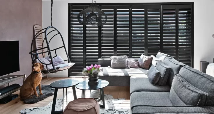 Woonruimte met een krijze l-vormige bank, een hangstoelm ronder bijzettafels en donkerbruine shutters voor de ramen